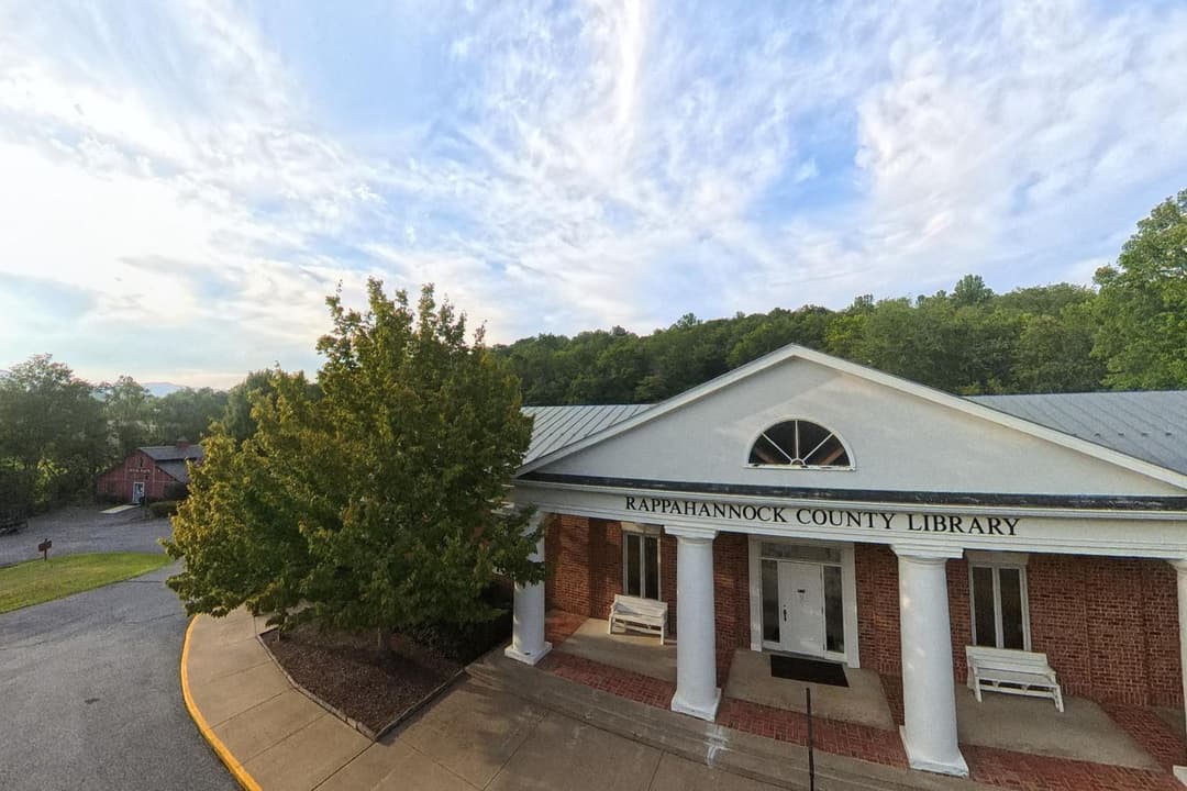 The Rappahannock County Library at sunset, a brick building with a white-columned portico, with the Book Barn visible beyond the trees to the left.